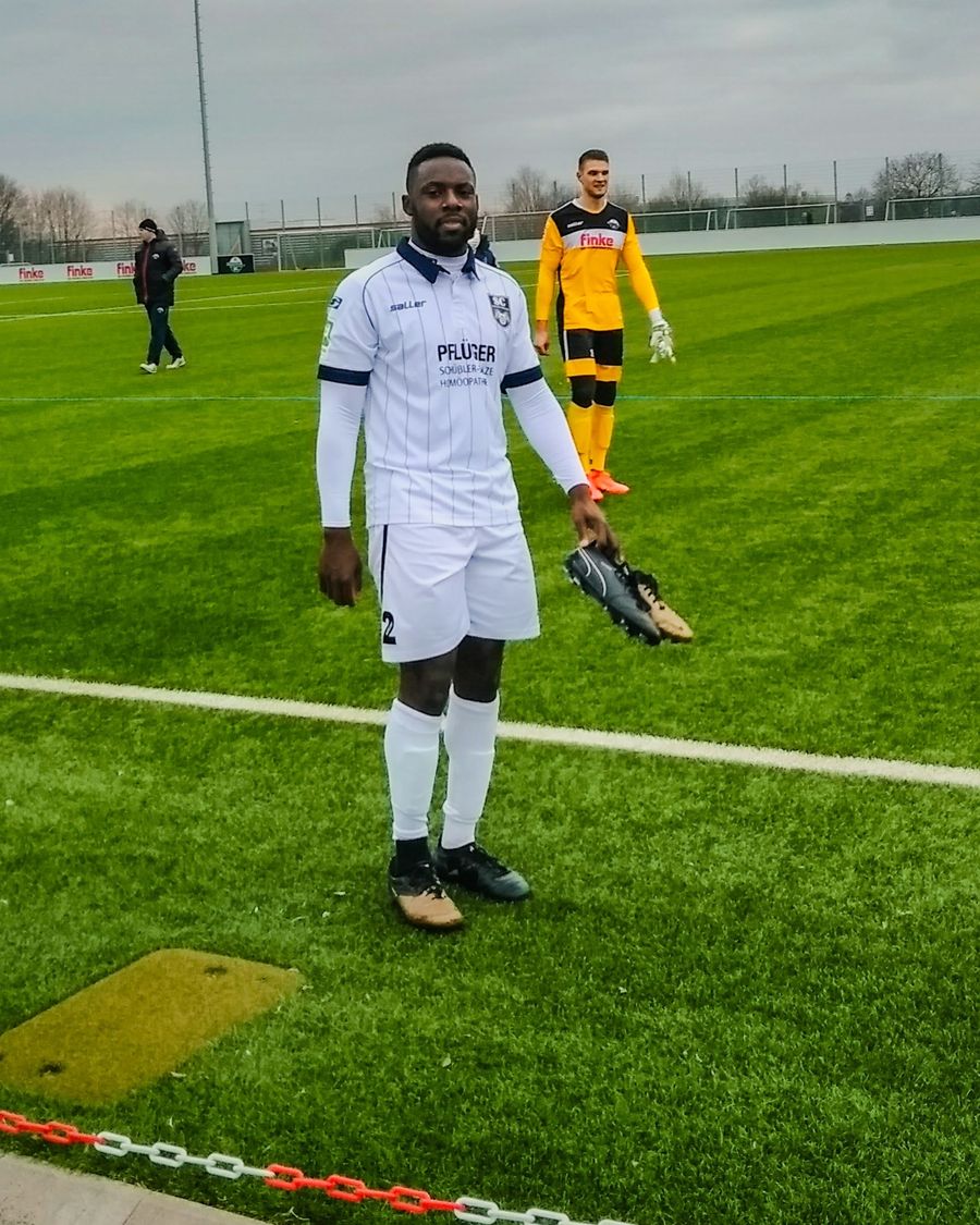 David Abunaw in a white match kit, boots in hand, at the side of a pitch in Germany